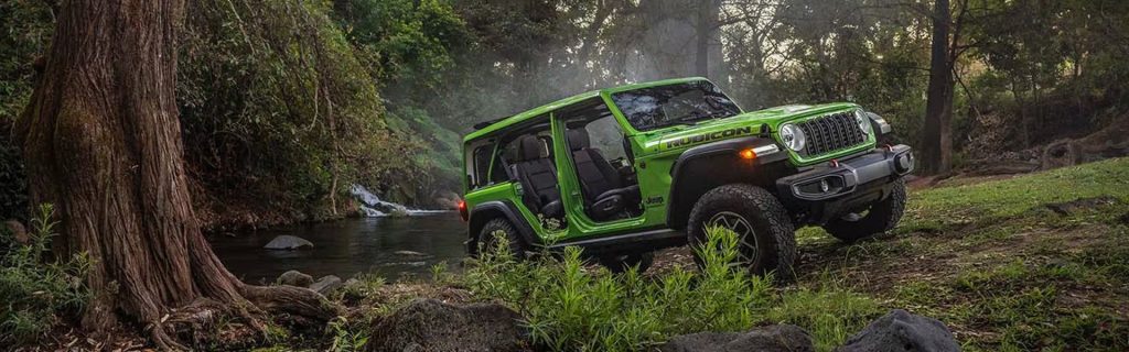 Green Jeep Wrangler driving off-road near a forest stream.
