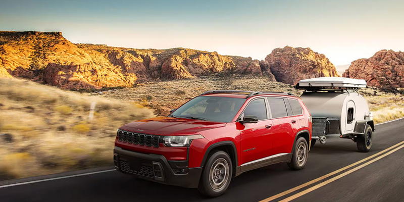 A red 2026 Jeep Grand Cherokee towing a small travel trailer on a scenic road surrounded by rocky terrain.