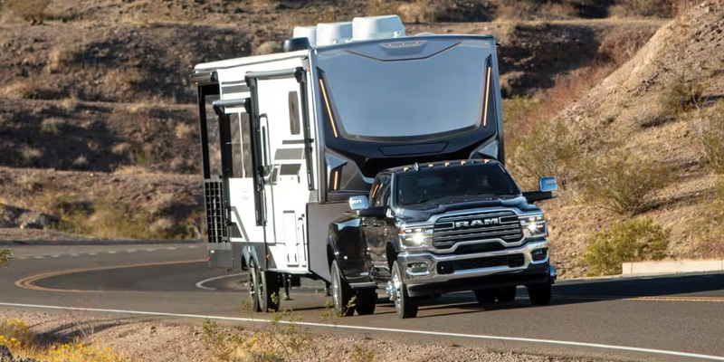 A 2026 RAM truck towing a large travel trailer on a curved road through a desert landscape.